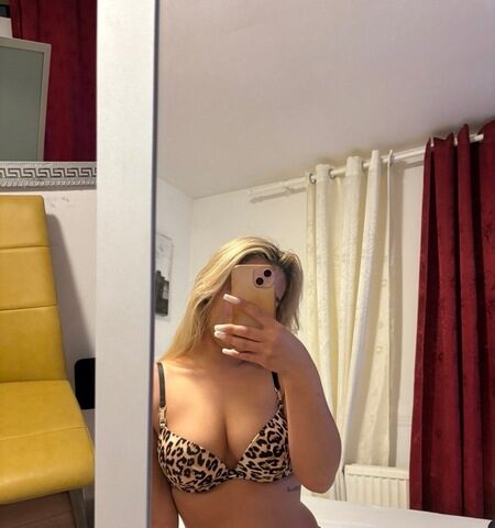 Eine Person in einem Bikini mit Leopardenmuster macht ein Spiegel-Selfie in einem Schlafzimmer mit roten Vorhängen und einem gelben Stuhl im Hintergrund.