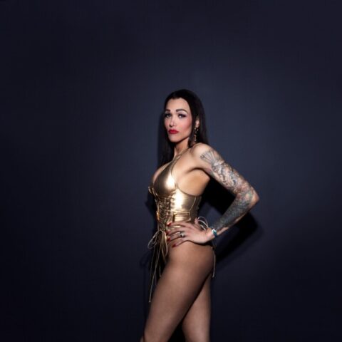 Frau in goldenem Korsett und Stiefeln posiert vor einem dunklen Hintergrund und zeigt ihre Tattoos.