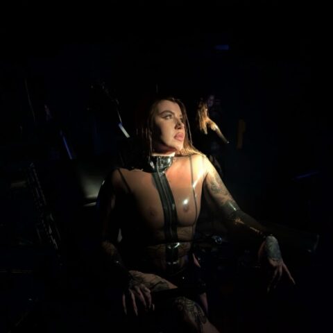 Eine Person, die ein glänzendes Ledergeschirr und ein Halsband trägt, sitzt in einem dunklen Raum, der von einem schmalen Lichtstrahl beleuchtet wird.