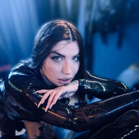 Eine Person in einem schwarzen Latex-Outfit posiert auf einem schwarzen gepolsterten Stuhl und blickt in einem schwach beleuchteten Raum mit einer Hand auf dem Kinn in die Kamera.