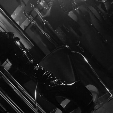 Ein hochhackiger Plateaustiefel sitzt auf einem modernen Metallstuhl in einem schwach beleuchteten Raum, in dem im Hintergrund Geräte zu sehen sind.