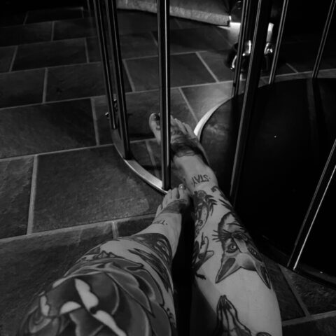 Schwarz-Weiß-Foto von tätowierten Beinen, die auf einem Steinboden ausgestreckt sind, mit Metallstäben und einem Teil einer runden Struktur im Hintergrund.