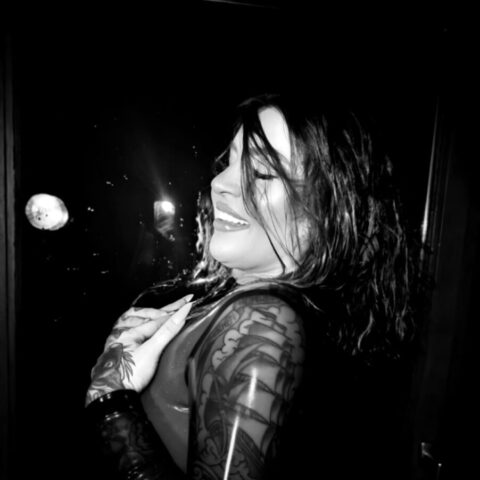 Eine Frau mit nassen Haaren und tätowierten Armen lächelt, während sie in einer schwach beleuchteten Umgebung von der Kamera wegschaut.