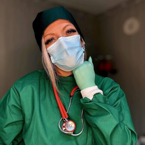 Person mit grünem Arztkittel, schwarzer Haube, Gesichtsmaske und Stethoskop, die eine behandschuhte Hand unter dem Kinn ablegt.