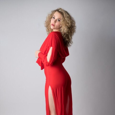 Frau mit lockigem blondem Haar steht seitwärts in einem roten Kleid mit hohem Schlitz und passenden roten Absätzen und posiert vor einem schlichten hellgrauen Hintergrund.