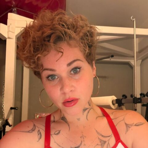 Eine Person mit kurzen lockigen Haaren und Ohrringen, die ein weißes Oberteil mit einem roten Kreuz trägt, steht in einem Raum mit sichtbaren Tätowierungen auf den Schultern und der Brust.