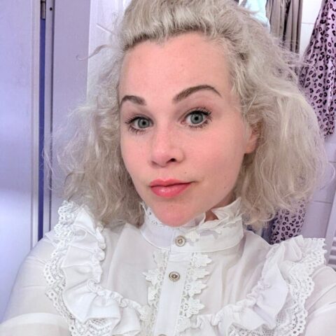 Eine Person mit hellen lockigen Haaren und weißer Rüschenbluse macht ein Selfie in einem Haus mit einem neutralen Gesichtsausdruck.