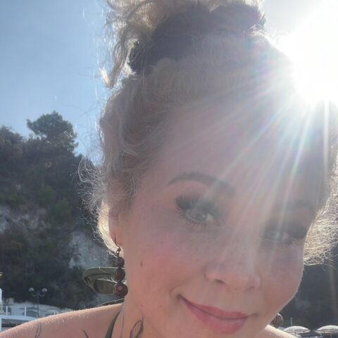 Eine Frau mit lockigem blondem Haar und Blumentattoos auf den Schultern lächelt an einem sonnigen Tag im Freien in die Kamera, während hinter ihr die Sonne scheint.