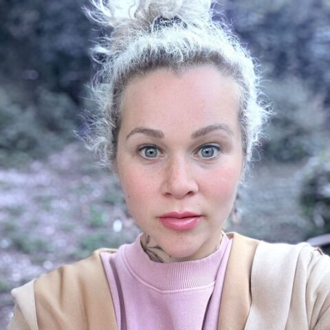 Eine Person mit hellem Haar in einem unordentlichen Dutt, die einen hellbraunen Mantel über einem rosafarbenen Oberteil trägt, blickt mit neutralem Gesichtsausdruck direkt in die Kamera; im Hintergrund ist unscharfes Grün zu sehen.