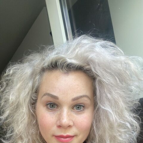 Eine Person mit lockigem, platinblondem Haar und sichtbaren Tätowierungen auf den Schultern trägt ein ärmelloses schwarzes Oberteil und schaut in die Kamera, während sie in einem Innenraum in der Nähe eines Fensters steht.