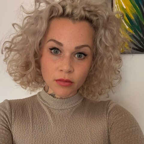 Person mit lockigem blondem Haar, hellen Augen und neutralem Gesichtsausdruck, die ein beiges Oberteil trägt, posiert in einem Innenraum vor einer weißen Wand, an der ein Teil eines bunten Gemäldes zu sehen ist.