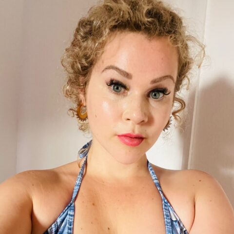 Eine Frau mit lockigem blondem Haar und blauen Augen blickt in einem gut beleuchteten Innenraum in die Kamera. Sie trägt ein blaues Neckholder-Top und Ohrringe.