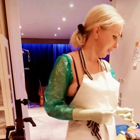 Eine Frau mit weißer Schürze, grünen Handschuhen und Schutzärmeln steht in einem gut beleuchteten Raum, in dessen Nähe medizinische oder Laborgeräte zu sehen sind.