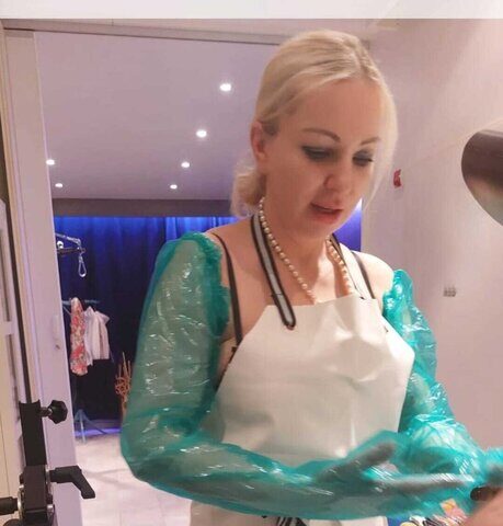 Eine Frau, die eine weiße Schürze, Plastikarmstulpen und eine Perlenkette trägt, steht in einem Haus und scheint sich auf eine Aufgabe vorzubereiten.