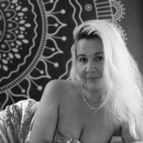 Eine Frau mit langen hellen Haaren liegt auf der Seite und trägt ein trägerloses Oberteil vor einer gemusterten Wand. Das Bild ist in schwarz-weiß gehalten.