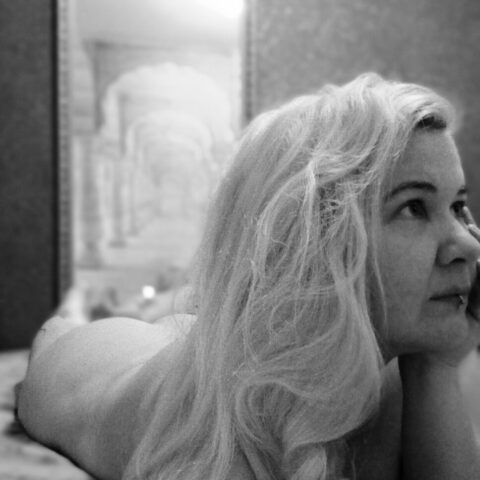 Eine Person mit langen blonden Haaren liegt auf dem Bauch auf einem Bett und schaut nach rechts. Das Bild ist in Schwarz-Weiß mit einem unscharfen Hintergrund.