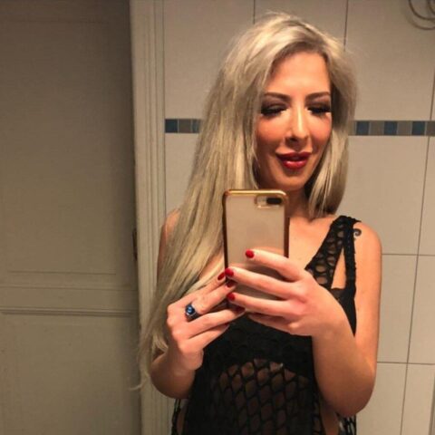 Eine Person mit langen blonden Haaren macht in einem Badezimmer ein Spiegel-Selfie. Sie hält ein Smartphone mit einer goldenen Hülle und trägt ein schwarzes Outfit.