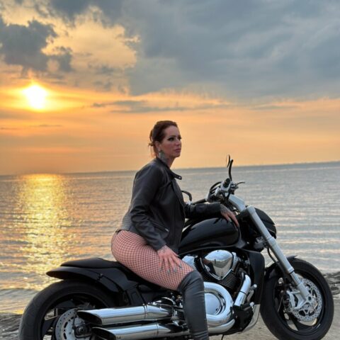 Eine Frau sitzt bei Sonnenuntergang auf einem schwarzen Motorrad in der Nähe einer Uferpromenade. Sie trägt eine Jacke, Netzstrümpfe und hochhackige Stiefel. Der Himmel ist teilweise bewölkt und die Sonne spiegelt sich im Wasser.