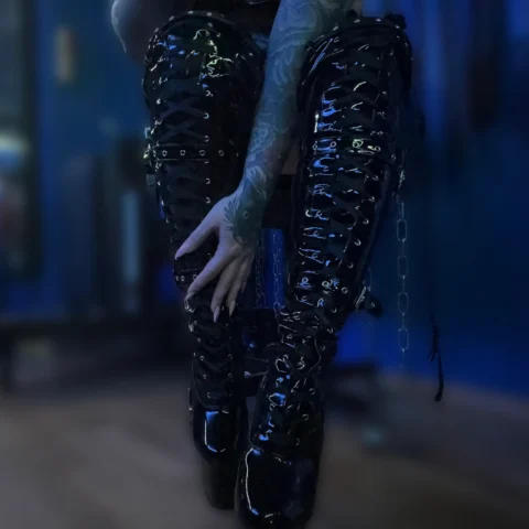 Person in glänzenden schwarzen Schnürstiefeln und mit tätowierten Armen, die auf einem Stuhl in einem schwach beleuchteten Raum mit Blautönen sitzt.