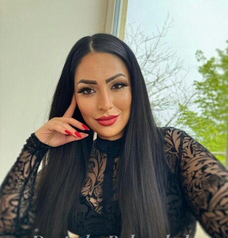 Frau mit langen schwarzen Haaren und auffälligem Make-up in einem schwarzen Spitzenoutfit sitzt drinnen am Fenster. Der Text auf dem Bild lautet „Deutsche Rubenslady Sensual Serie“.