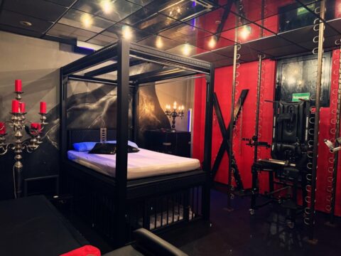 Ein schwach beleuchteter Raum mit einem Himmelbett, roten und schwarzen Wänden, einer großen X-förmigen Struktur, Fitnessgeräten und Kerzenhaltern als Dekoration.