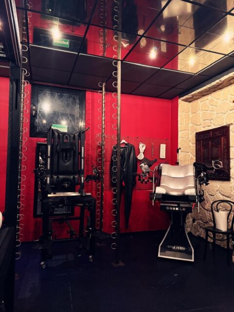 Ein Raum mit roten Wänden, einer verspiegelten Decke, einer Metallstange, zwei BDSM-Stühlen, hängenden Fesseln, einem schwarzen Bodysuit und einem Holzstuhl.