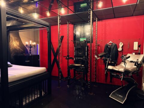 Ein Raum mit BDSM-Möbeln, darunter ein Himmelbett, ein Andreaskreuz, ein medizinischer Stuhl, Lederfesseln und verschiedene Ausrüstungsgegenstände vor roten und schwarzen Wänden.