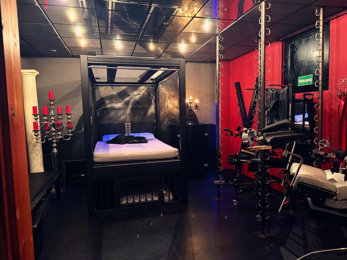 Ein schwach beleuchteter Raum mit einem großen Himmelbett, roten und schwarzen Wänden, Kerzenhaltern und verschiedenen Übungs- und Fesselungsgeräten.