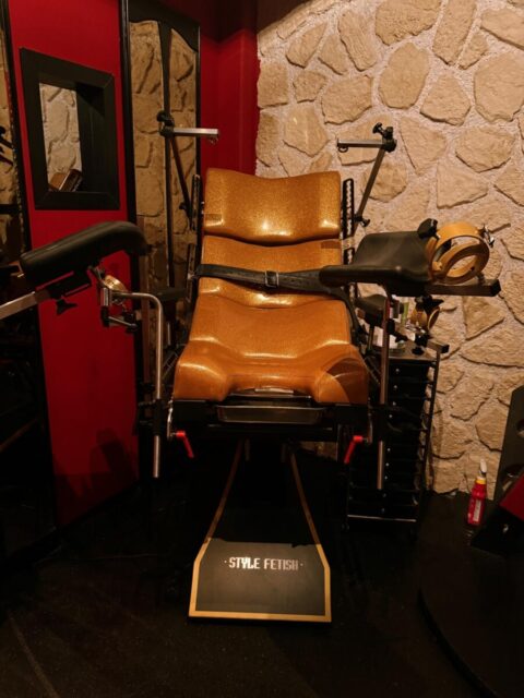 Ein goldener Lehnstuhl mit Fesseln und verschiedenen Aufsätzen steht in einem Raum mit steingemusterten Wänden und roten Akzenten. Auf einem Schild am Sockel steht "STYLE FETISH".