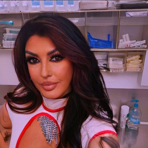 Eine Frau mit langem dunklem Haar und auffälligem Make-up macht in einer medizinischen Umgebung ein Selfie. Sie steht vor Regalen mit medizinischem Zubehör und einem Spender für Händedesinfektionsmittel.