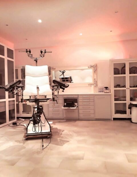 Ein medizinischer Untersuchungsraum mit einem leeren gynäkologischen Untersuchungsstuhl, medizinischen Geräten, Schränken und warmer Beleuchtung.