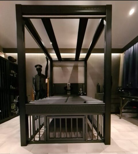 Ein großes, schwarzes Himmelbett aus Metall mit einem Käfig darunter steht in einem schwach beleuchteten Raum mit dunklem Mobiliar und einer Schaufensterpuppe in Uniform im Hintergrund.