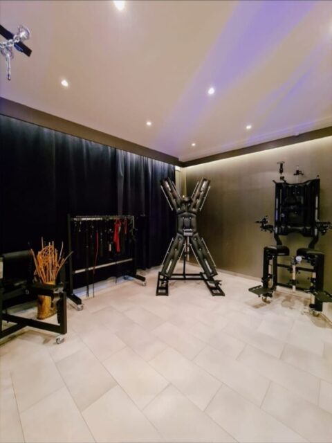 Ein geräumiger Raum mit gefliestem Boden ist mit BDSM-Möbeln ausgestattet, darunter ein Andreaskreuz, ein Bondage-Stuhl, Fesseln und verschiedene Accessoires vor dunklen Wänden und einem Vorhang.