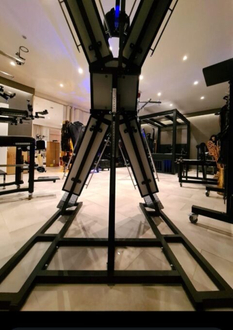 Ein großer X-förmiger Metallrahmen steht in der Mitte eines gut beleuchteten Raums, der mit verschiedenen Geräten und Maschinen gefüllt ist.