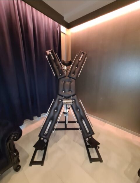 Ein großer, gepolsterter, X-förmiger Fesselungsrahmen steht aufrecht in einem Raum mit dunklen Vorhängen und einer grauen Wand.