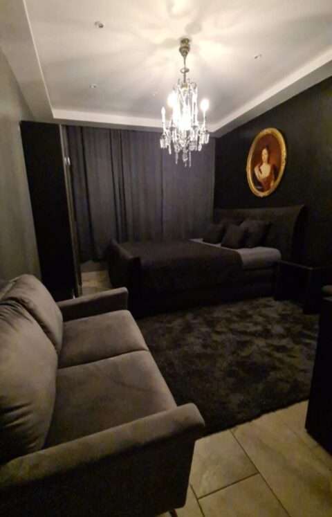 Ein schwach beleuchtetes Schlafzimmer mit schwarzen Wänden, einem Bett, einem grauen Sofa, einem dunklen Teppich, einem Kronleuchter, geschlossenen Vorhängen und einem gerahmten Porträt an der Wand.