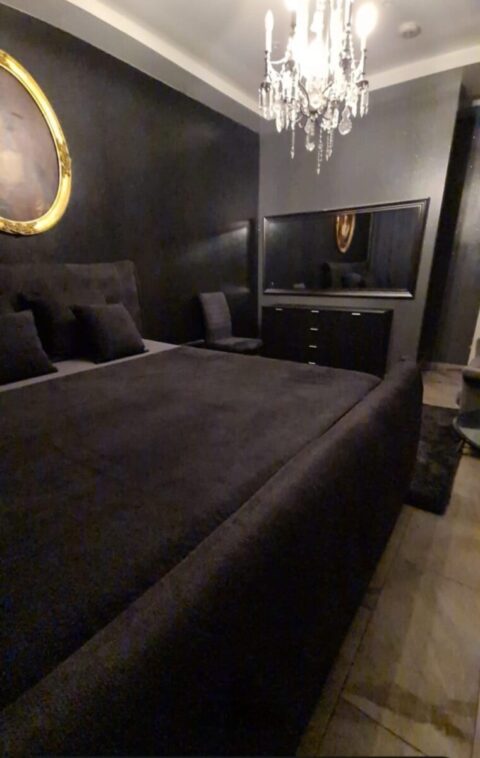 Ein Schlafzimmer mit schwarzen Wänden und Möbeln, einem Kronleuchter, einem ovalen, goldgerahmten Kunstwerk, einem großen Spiegel und einem Bett mit schwarzem Bettzeug und Kissen.