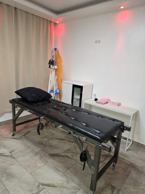 Ein Raum mit einem gepolsterten schwarzen Untersuchungstisch mit Fesseln, einem Kissen, medizinischen Geräten, gefalteten Handtüchern und sanfter roter Beleuchtung.