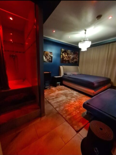 Ein schwach beleuchtetes Schlafzimmer mit einem großen Bett, einem Kronleuchter, Wandkunst und einem rot beleuchteten Duschbereich auf der linken Seite.