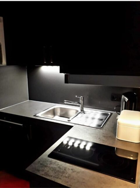 Moderne Küche mit einer Edelstahlspüle, einem Elektrokochfeld, einem Wasserkocher und einem weißen Vorratsbehälter auf einer grauen Arbeitsplatte mit Schrankbeleuchtung.