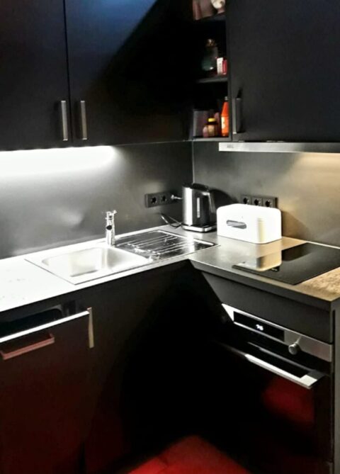 Moderne schwarze Küche mit Spüle, Herd, Backofen, Toaster, Wasserkocher und Unterschrankbeleuchtung; auf der Arbeitsplatte und in den Regalen befinden sich verschiedene Kleinigkeiten.