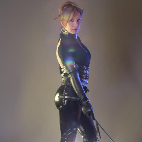 Eine Person in einem schwarzen Latex-Outfit, High Heels und Handschuhen hält eine Reitgerte in der Hand. Helles Licht leuchtet von hinten und erzeugt einen dramatischen Silhouetteneffekt.