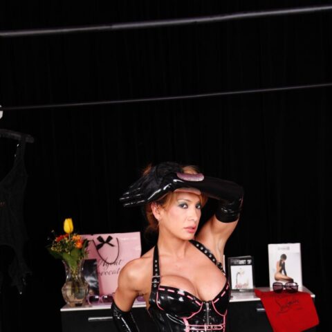 Eine Frau in einem schwarz-rosa Latex-Outfit posiert drinnen mit einer Hand an ihrem Hut. Im Hintergrund sind verschiedene Wäscheartikel, Blumen und Schachteln zu sehen.