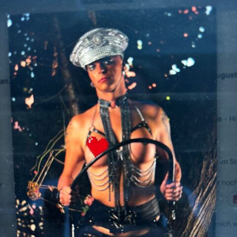 Eine Person, die einen Metallhut, Kettenzubehör und freizügige Kleidung trägt, steht nachts im Freien und hält eine Reitgerte.