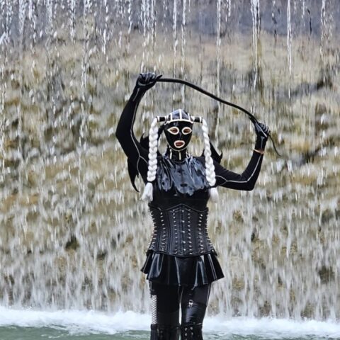 Eine Person in schwarzem Latex-Outfit, Korsett und Maske steht an einem felsigen Ufer und hält eine Peitsche in der Hand, im Hintergrund ein Wasserfall.