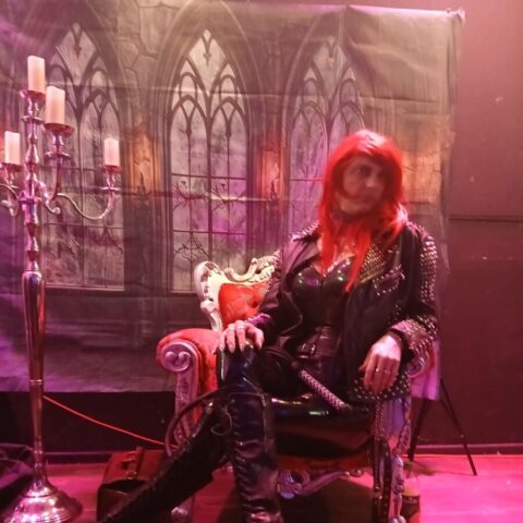 Eine Person mit langen roten Haaren, die ein schwarzes Nietenkostüm und Plateaustiefel trägt, sitzt auf einem verzierten Stuhl vor einer Kulisse im gotischen Stil und einem Kerzenleuchter.