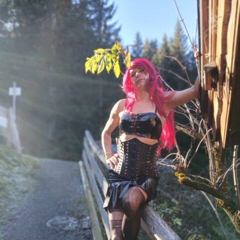 Eine Person mit langen rosafarbenen Haaren, die ein schwarzes Vinyl-Outfit und Strümpfe trägt, posiert an einem Holzzaun entlang eines Waldwegs im Sonnenlicht.