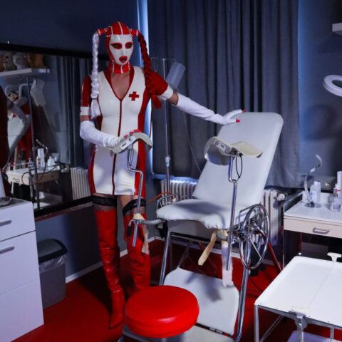 Eine Person in einem rot-weißen, an eine Krankenschwester erinnernden Latex-Outfit und einer Maske steht neben einem medizinischen Untersuchungsstuhl in einem schwach beleuchteten klinischen Raum.