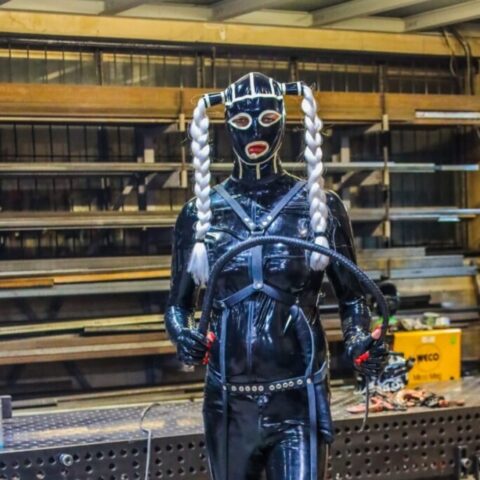 Eine Person in einem schwarzen Ganzkörper-Latexanzug mit Maske, weißen geflochtenen Zöpfen und hohen Stiefeln steht in einer Industriehalle und hält eine Peitsche.