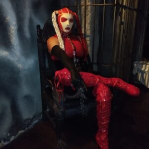 Eine Person mit einer dramatischen rot-weißen Maske, einem rot-schwarzen Kostüm und oberschenkelhohen, glänzenden roten Stiefeln sitzt auf einem dunklen Holzstuhl in einem schwach beleuchteten Raum mit blau-grauen, strukturierten Wänden.